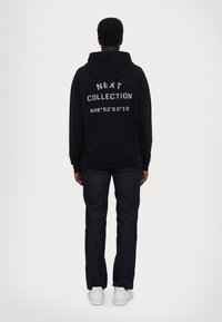 Osoba zády k nám, oblečená v černé mikině s kapucí a nápisem "NEXT COLLECTION N48°52'E2°18", tmavých džínách a bílých teniskách na jednobarevném pozadí.