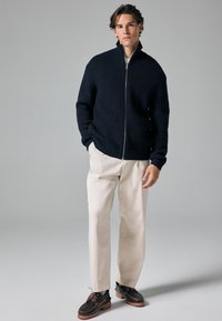 Marine geribbelde zip-up trui, crèmekleurige broek en donkere schoenen. De trui heeft een hoge kraag, een ontspannen pasvorm en een stevige stof.
