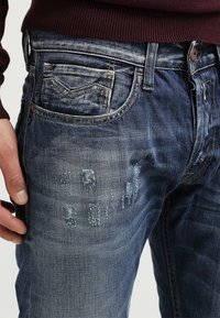Jeans de mezclilla con un color azul desgastado, parches con desgaste y un diseño clásico de cinco bolsillos con cierre de botón en la cintura.