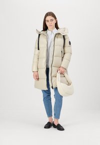 Beige puffer coat with a hood, zip closure, and black straps. Worn over a white shirt and denim, paired with black loafers and a light bag.