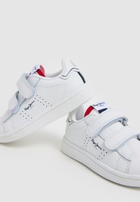 Valged tennisekingad siledast nahast viimistlusega, millel on kaks Velcro rihma, aukudega detailid ja logo silt keelel.
