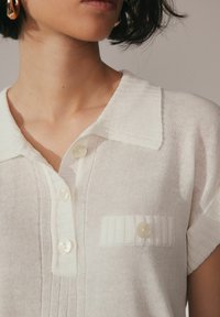 Valge kudumisega polo särk, millel on laiale haardele mõeldud krae, kolm läbipaistvat nuppu ja tekstuuriga tasku detail vasakul rinnal. Pehme kangas.