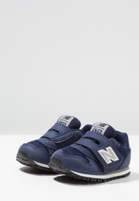 Tmavě modré tenisky z meshové a semišové látky, s pásky na suchý zip, polstrovaným jazykem a bílým logem New Balance na straně.