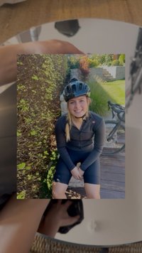 Smaidoša sieviete melnā velosipēda aprīkojumā un ķiverē sēž uz koka klāja blakus dzīvžogam, kamēr fona redzams velosipēds, kas atbalstīts pret akmens pakāpieniem.