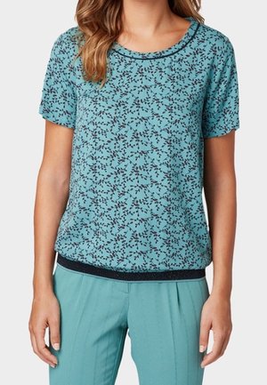 Femme portant un haut à manches courtes turquoise avec un imprimé floral foncé et un pantalon turquoise assorti, debout les mains détendues le long du corps.