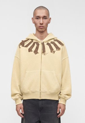 Jeune homme portant un sweat à capuche beige zippé avec de grandes lettres marron foncé usées sur les épaules, debout devant un fond blanc uni.