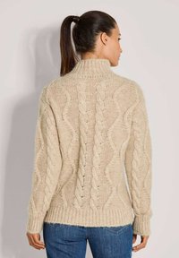 Frau mit einem hohen Pferdeschwanz, die einen cremefarbenen Kabelstrickpullover und eine blue Jeans trägt, mit dem Rücken zu einer schlichten hellen Hintergrund.