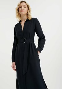 Vestido negro con cinturón que presenta un cuello con solapa, mangas largas con puños y aberturas laterales para facilitar el movimiento. Textura suave.