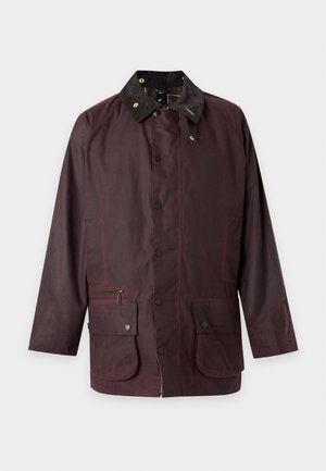 Homme portant une casquette marron, une veste marron foncé, une chemise à carreaux verts et noirs, une ceinture marron, un pantalon beige et des chaussures marron, debout devant un fond uni.