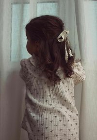 Lysefarvet, mønstret bluse med flæser ved halsen; bomuldsstof; barn med krøllet hår bundet op med et hvidt bånd, der kigger ud af et vindue.