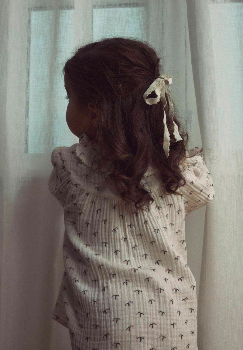 Lysefarvet, mønstret bluse med flæser ved halsen; bomuldsstof; barn med krøllet hår bundet op med et hvidt bånd, der kigger ud af et vindue.