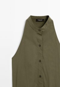 Blouse sans manches vert olive avec un col haut, présentant un plastron à boutons et une texture de tissu lisse. Étiquette de la marque visible au niveau du col.