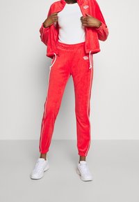 Röd velourbyxdräkt med vita detaljer, bestående av en jacka med dragkedja och byxor med dragsko, matchad med vita sneakers.