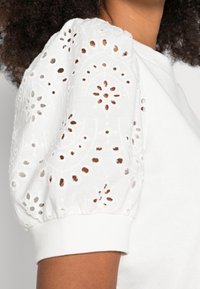 White puff sleeve with embroidered eyelet floral patterns on a person wearing a white garment, showing shoulder and upper arm.