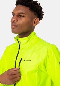 Veste coupe-vent jaune néon avec fermeture éclair à l'avant, col haut et détails de logo. Caractérisée par un matériau léger et texturé.