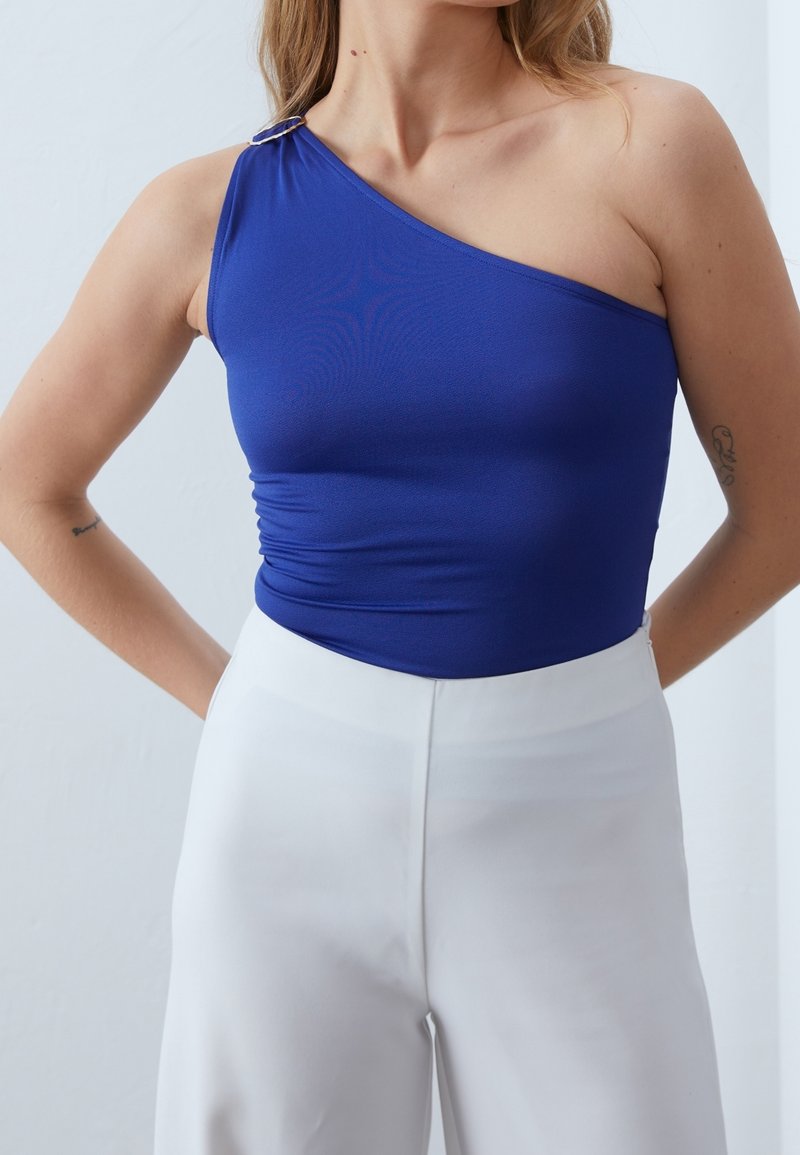 Kungligt blå enaxlad bodysuit med rynkat sidofåll och silverdetaljer, kombinerad med vita högmidjade byxor i en slät textur.