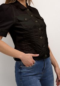 Svart kortärmad blus med korta puffärmar, knäppning i fronten, två bröstfickor och en strukturerad silhuett, stylad med blå jeans.