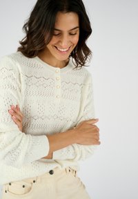 Pull en laine blanc tricoté avec un motif en losange texturé, décolleté rond et détails de boutons sur le devant. Matériau doux et léger.