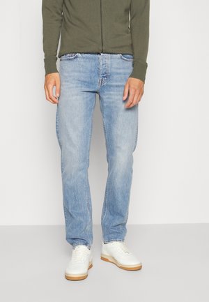 Man draagt lichtblauwe jeans, witte sneakers en een olijfgroene longsleeve, staand tegen een effen achtergrond.