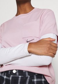 Rosa t-shirt med rund halsringning och bröstficka. Har en liten silverfärgad Nike-logga på framsidan. Lager på lager med långa vita ärmar under.