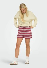 Shorts bordeaux avec des rayures horizontales blanches, taille élastique avec un cordon de serrage, associés à un sweat-shirt crème et des chaussures blanches.