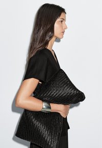 Svart vävd kuvertväska hålls nära, vilket visar en texturerad yta. Modellen bär en enkel svart topp och ett silverfärgat armband.