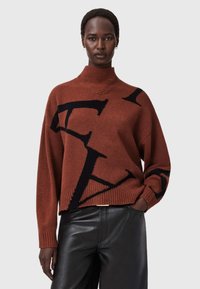 Brązowy sweter robiony na drutach z wysokim kołnierzem, zdobiony dużymi czarnymi literami logo na przodzie. Żebrowane mankiety i dół. Nosić go na czarnych skórzanych spodniach.