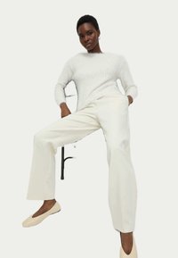 Pull en maille blanc, pantalon large crème et chaussures slip-on beige. Le modèle pose avec un bras sur la chaise, affichant une coupe décontractée.