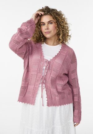Donna con capelli ricci che indossa un cardigan rosa a maglia aperta annodato davanti sopra un vestito bianco, che si tocca i capelli con una mano.