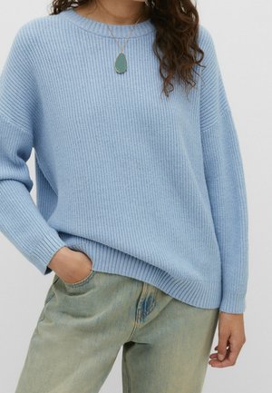 Mujer con suéter azul claro acanalado, vaqueros azul verdoso, con una mano en el bolsillo y un collar con colgante en forma de lágrima.