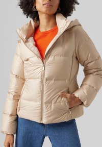 Beige-täytteinen puffer-takki, jossa on vetoketju edessä, kiinnitetty huppu ja sivutaskut; tunnistaa tikatun rakenteen ja kultakullanväriset vetoketjun yksityiskohdat.