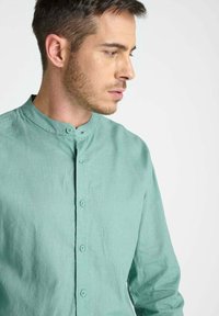 Jonge man met kort haar in een lichtgroen knoopshirt met een Mandarin-kraag, die naar rechts kijkt tegen een effen achtergrond.