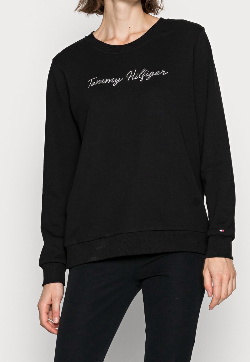 Personne portant un sweat-shirt noir Tommy Hilfiger avec un logo en écriture cursive blanche sur la poitrine et un petit logo drapeau sur le poignet de la manche.