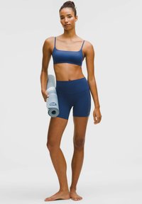 Vrouw staat blootsvoets in een marineblauwe sportbeha en shorts, met een opgerolde lichtblauwe yogamat tegen een effen achtergrond.