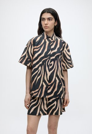 Kobieta o długich ciemnych włosach, ubrana w beżową i czarną koszulkę w paski zebry z krótkim rękawem oraz pasujące szorty, stojąca na jednolitym tle.