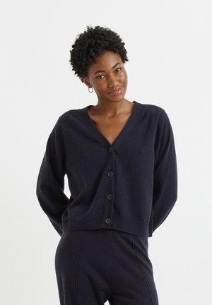 Vrouw met kort krullend haar, een donker marineblauw gestoken vest en bijpassende broek, staand met de handen achter de rug tegen een witte achtergrond.