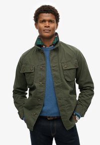 Groene stoffen jas met een staande kraag en voorzakken, gedragen over een blauwe textuurtrui en een geruite laagshirt. Donkere jeans zichtbaar.