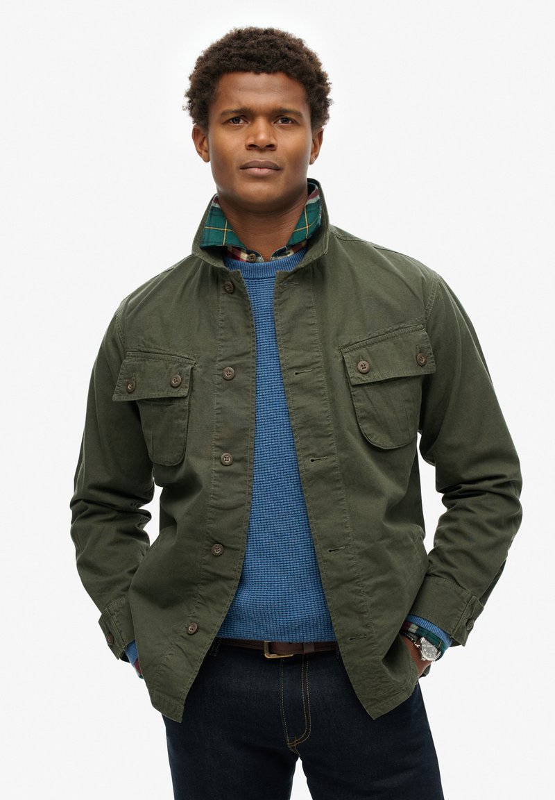Groene stoffen jas met een staande kraag en voorzakken, gedragen over een blauwe textuurtrui en een geruite laagshirt. Donkere jeans zichtbaar.