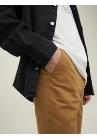 Personne portant un pantalon beige avec une main dans la poche, une chemise blanche partiellement rentrée et une veste noire boutonnée avec les manches baissées.