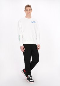 Felpa bianca a maniche lunghe con logo blu e verde sul petto e sulla manica, abbinata a pantaloni neri e sneaker nere, rosse e bianche.