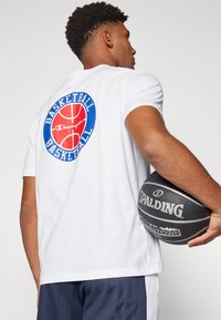 T-shirt Champion blanche avec un grand graphique de basketball rouge et bleu au dos, associée à un short de sport marine et un ballon de basketball noir.