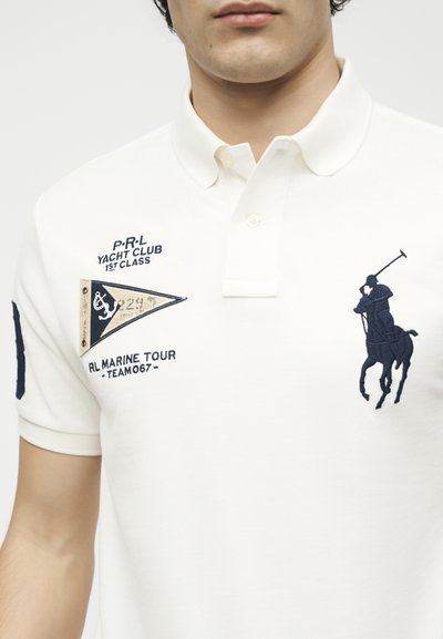 Λευκό πόλο μπλουζάκι με κεντημένο σε σκούρο μπλε παίκτη πόλο, κείμενο yacht club και τριγωνικό σήμα σημαιάκι στο επάνω μέρος του στήθους.