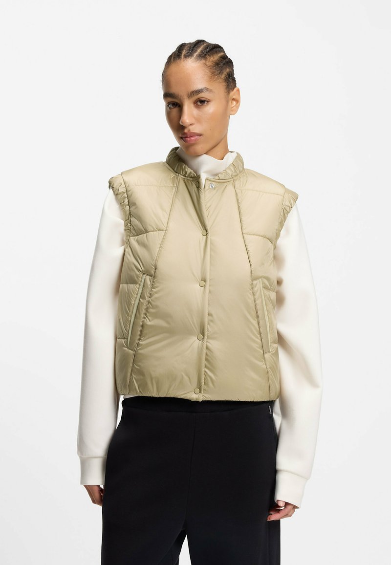 Frau mit geflochtenem Haar, die eine beige gesteppte, ärmellose Weste über einem weißen Langarmshirt und schwarze Hose vor einem einfarbigen Hintergrund trägt.