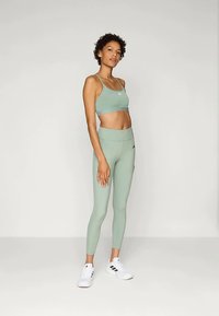 Γυναίκα που στέκεται σε ανοιχτό πράσινο αθλητικό σουτιέν και κολάν, φορώντας λευκά αθλητικά παπούτσια, σε απλή ανοιχτή φόντο.