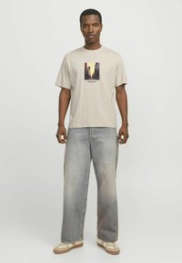 Beige bomull t-skjorte med grafisk trykk av en bylandskap i midten. Brukt sammen med løse, falmede denimjeans og beige joggesko.