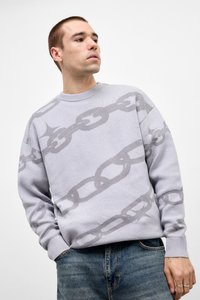 Ung mand iført en lys grå sweater med et stort kædemønster, står med den ene hånd i sin jeanslomme og kigger til siden.
