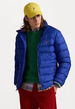 Doudoune bleue avec un design matelassé, associée à un pull en maille vert, une chemise à carreaux, un pantalon en velours côtelé rouge et une casquette jaune.
