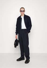 Schwarze Bomberjacke, weißes T-Shirt und marineblaue Hose. Schwarze Sonnenbrille und Schuhe. Hält einen schwarzen Rucksack. Schlichtes, reduziertes Design.