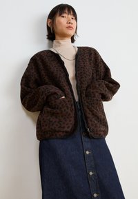 Smeđa jakna od pliša s uzorkom leoparda, s patentnim zatvaračem, oversized oblikom i prednjim džepovima, u kombinaciji s traper suknjom koja ima detalje s gumbićima.