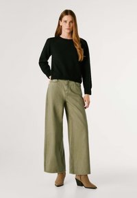 Melnā adīta džempera, plata olīvu zaļa džinsa bikses un gaiši brūnas augstpapēžu zābaki līdz potītei. Džemperim ir apaļa kakla izgriešana; biksēm ir brīva piegriezuma stils ar priekšējiem kabatām.